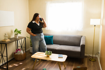 Full length view of a plus size woman relaxing at home, drinking coffee in living room, thinking and enjoying free time