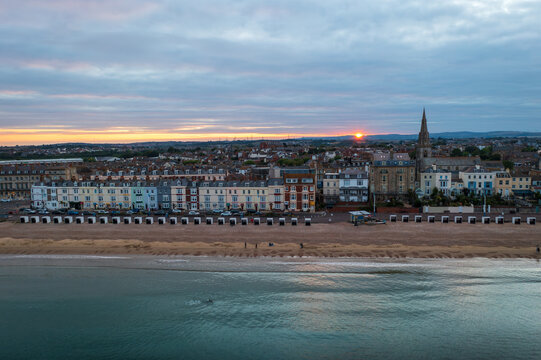 Aerial view of the golden sun kissing the horizon over the seafront and spire of Holy Trinity Church, casting a warm glow on Weymouth's sandy beach, Weymouth, England, United Kingdom.