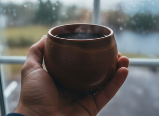 Man holding a rustic ceramic cup of steaming hot coffee or tea by a window on a rainy day, for cozy morning concept.