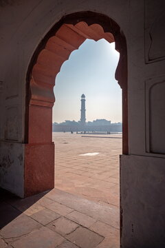 View of the stark minaret piercing the hazy sky through the archway's embrace, casting shadows on the ancient ground, Lahore, Badshahi Masjid Mosque, Pakistan.