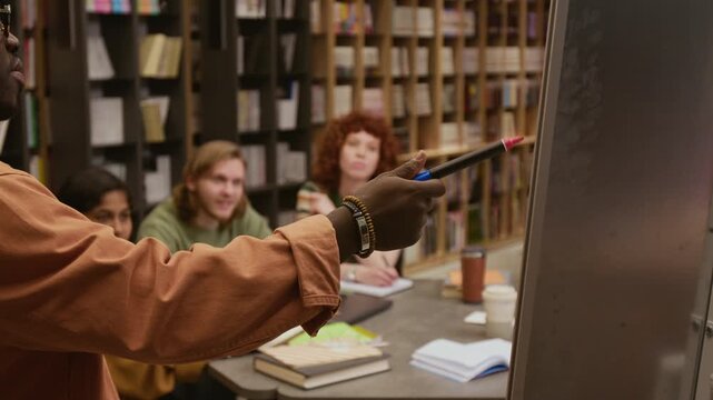 Cropped shot of African American male teacher pointing with marker at whiteboard presenting new topic to multi ethnic group of adult students sitting at desk in library