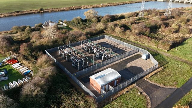 Aerial view of an electrical substation nestled amongst autumnal foliage near a winding river, its metallic structures gleaming against the muted landscape, Kings Lynn, United Kingdom.
