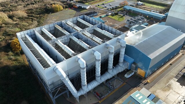 Aerial view of a large industrial cooling tower with steam rising against a backdrop of buildings and trees, Kings Lynn, United Kingdom.