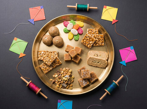 Assorted makarsankranti sweets arranged in a brass plate with real kites and manja decor around it on a premium matte charcoal grey background in clean top view festive style