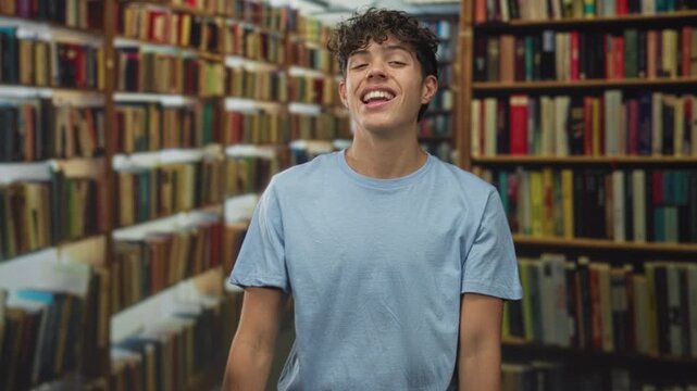 Boy in blue shirt pinches fingers with hands, grinning and making a money gesture surrounded by tall bookshelves in a building library aisle; playful joy.