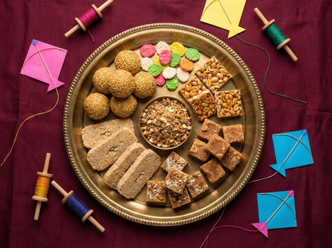 Brass plate filled with makarsankranti sweets and real kite festival decor arranged around the plate on a premium deep maroon matte background in clean top view festive composition