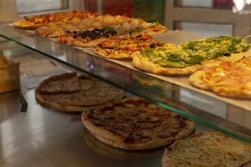 assorted rectangular pizzas on counter, warm glass display with pesto and basil accents, golden crusts and melted mozzarella
