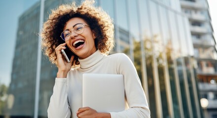 Woman laughing while talking on phone and holding a laptop by office glass facade