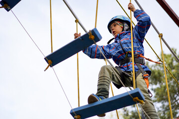 Low-angle shot of a young boy balancing on a suspended plank bridge high above the ground on a ropes course against a bright sky.