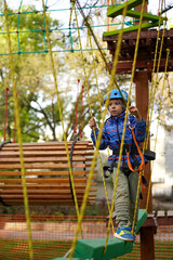 Young boy with helmet and harness carefully balances on a plank walk high above the ground on a ropes course.