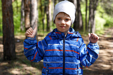 Portrait of a serious young boy in a blue jacket and white beanie giving a subtle gesture in a sunny, dappled forest. giving a thumbs up