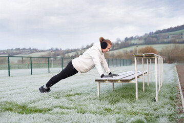 A woman in a white hoodie, gloves, and earmuffs trains a push-up on a  wooden benches on frosted field. This athletic outdoor scene highlights fitness, perseverance, and seasonal training
