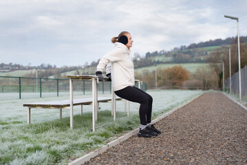 Woman exercising outdoors on frosty morning using park bench for tricep dips and leg workout. A woman feels both physically and emotionally better after training