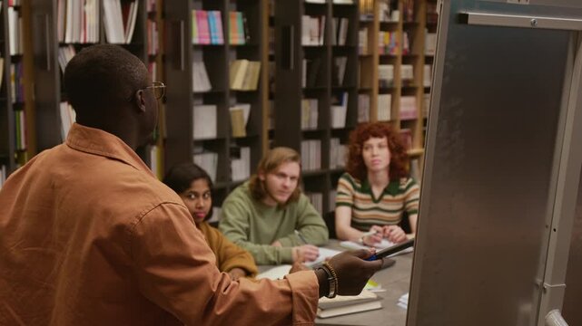 Rear view of African American male teacher pointing at whiteboard introducing lesson topic to smiling adult students sitting at desk in library during language training class
