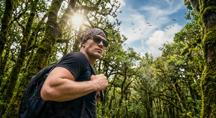 Low angle view of adventurous male hiker standing in ancient mossy forest looking up at tall trees and sunlight