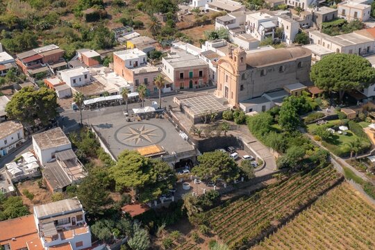 Aerial view of the church tower rising amidst the village buildings, contrasting with the circular plaza's compass design and nearby vineyards, Salina, Sicilia, Italy.