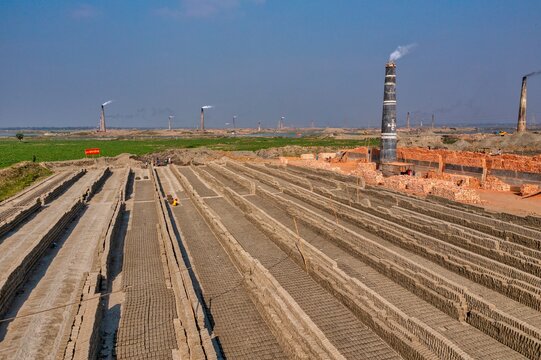 Aerial view of long rows of bricks drying under the clear sky, with distant chimneys releasing trails of smoke, Aminbazar, Dhaka Division, Bangladesh.