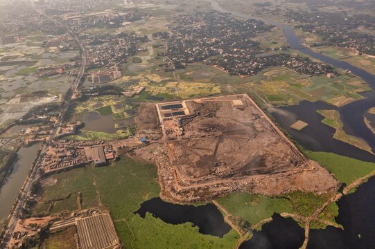 Aerial view of a sprawling landfill site juxtaposed against the meandering river and patches of vibrant green vegetation, Aminbazar, Dhaka Division, Bangladesh.