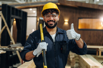 Big hammer is in hands. Portrait of a male carpenter posing in a woodworking workshop