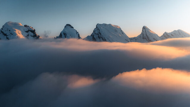 Aerial view of jagged, snow-capped peaks rise majestically above a sea of swirling clouds, illuminated by the warm glow of the rising sun, Wildhaus-Alt Saint Johann, St. Gallen, Switzerland. - Powered by Adobe