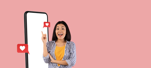 A young woman stands beside a tall smartphone screen, smiling and pointing with enthusiasm. She is excited about social media interactions, surrounded by heart icons that symbolize likes or love.