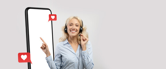 A cheerful woman with headphones is smiling and pointing at a large smartphone screen displaying empty space. She appears engaged and happy in a bright, neutral environment.