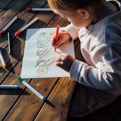 A young child is sitting at a wooden table and drawing on a piece of white paper with colorful markers.