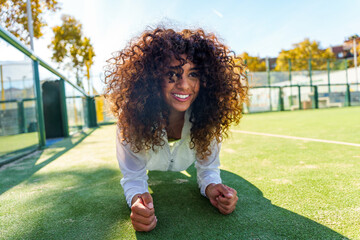 Young woman with curly hair smiling, doing plank exercise for fitness and core strength on outdoor court