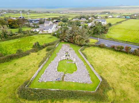 Aerial view of a triangular stone structure, nestled in vibrant green fields against a backdrop of distant houses and the sea, Carrowmore, County Sligo, Ireland.