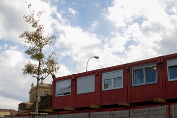red modular office containers under cloudy sky, stacked above wooden fence with shuttered windows, metal grill and street lamp,