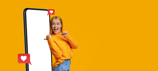 A young woman wearing a bright orange sweater smiles widely, showing excitement as she engages with a large smartphone.