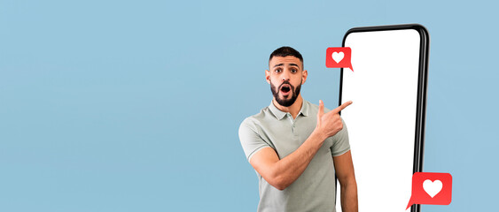 A man stands in front of a blue wall, pointing towards a large blank smartphone screen with surprise. Social media icons float next to him, showcasing excitement and engagement.