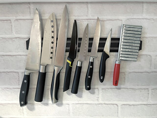 Collection of various kitchen knives displayed on a magnetic strip against a white brick wall, showcasing culinary tools for food preparation and cooking techniques