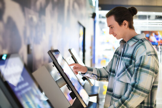 man using self-service kiosk with touchscreen. Guy selecting options on interactive terminal in cinema or mall.