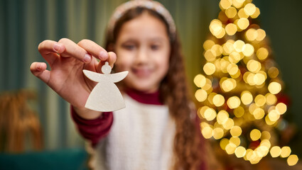 Smiling little girl holding a white angel Christmas ornament in front of glowing holiday tree lights, warm festive atmosphere, shallow depth of field and cozy indoor celebration mood