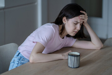 Unwell young woman with headache sits at table with coffee mug, touching forehead feeling exhausted after sleepless night. Fatigued female suffering head hurt, hangover in morning 