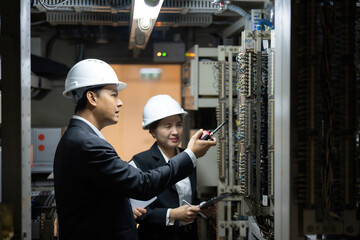 Asian male and female engineers inspect complex electrical panels inside an industrial control room, analyzing wiring connections and recording technical data for system optimization.
