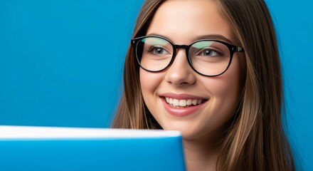 Young woman in glasses smiles, peering from behind a tablet on a blue backdrop, suitable for education, technology, and health.