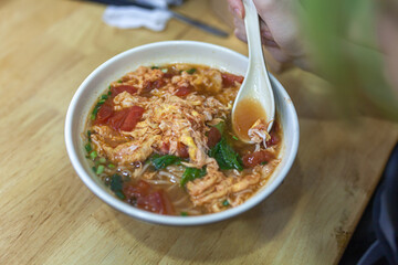Close-up of white ceramic bowl filled with tomato noodle soup topped with scrambled egg, chopped green onions and leafy greens on wooden table. Authentic coastal Chinese food with local ingredients.