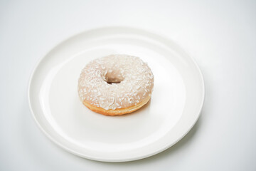 A donut with a smooth glaze and coconut flakes is placed on a white plate. The scene shows the treat ready for someone to take a bite. The light draws attention to the donut.