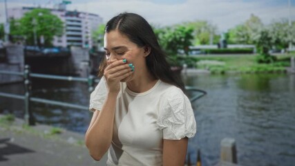 Young hispanic woman laughing with hand over mouth and turquoise nails on canal street in amsterdam; lighthearted amusement.