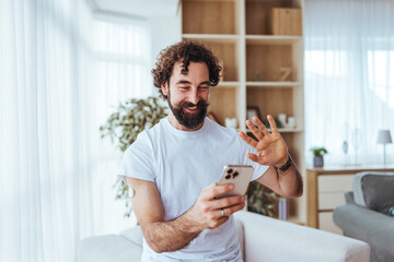 Happy Man Waving While Video Calling From Home With Smartphone in Cozy Living Room Today