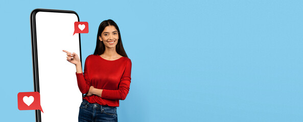 A joyful woman stands next to a large mobile phone with a blank screen, pointing at it. Bright blue...