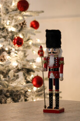 Wooden nutcracker in red uniform standing beside a softly lit tree decorated with red baubles.
