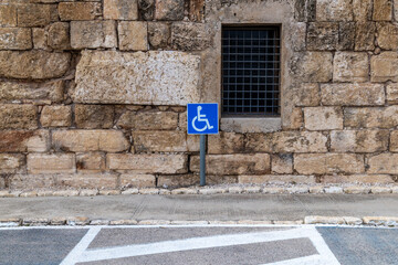 Accessible parking sign in a park against an old stone building wall, blue wheelchair symbol, designated disabled parking space, urban heritage setting, safety and accessibility concept