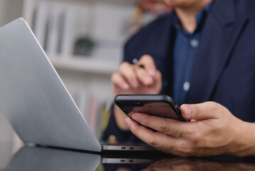 Close-up of a businessman's hands holding a smartphone and a pen while working on a laptop computer at an office desk. Concept of multitasking and modern business technology.