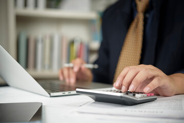 Close-up of an accountant's hands using a calculator to calculate tax or financial budget on a wooden desk with a laptop and documents.