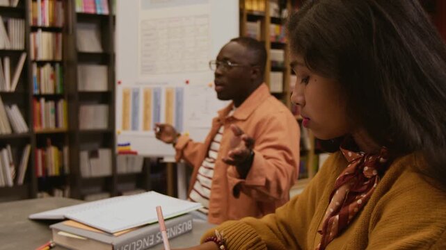 Side view of young South Asian woman writing in notebook during English group class while Black male teacher explaining irregular verbs forms pointing at whiteboard in library