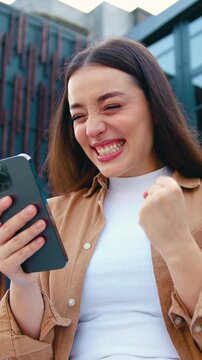 Close up of amazed girl holds smartphone, happy to receive notification, gestures actively from happiness, outdoors. Emotions and pleasant feelings concept