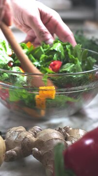 A vertical shot of a man mixing a fresh vegetarian salad in a glass bowl with a wooden spoon. Delicious and healthy food prepared with organic ingredients in a bright and cozy home kitchen. Close-up.